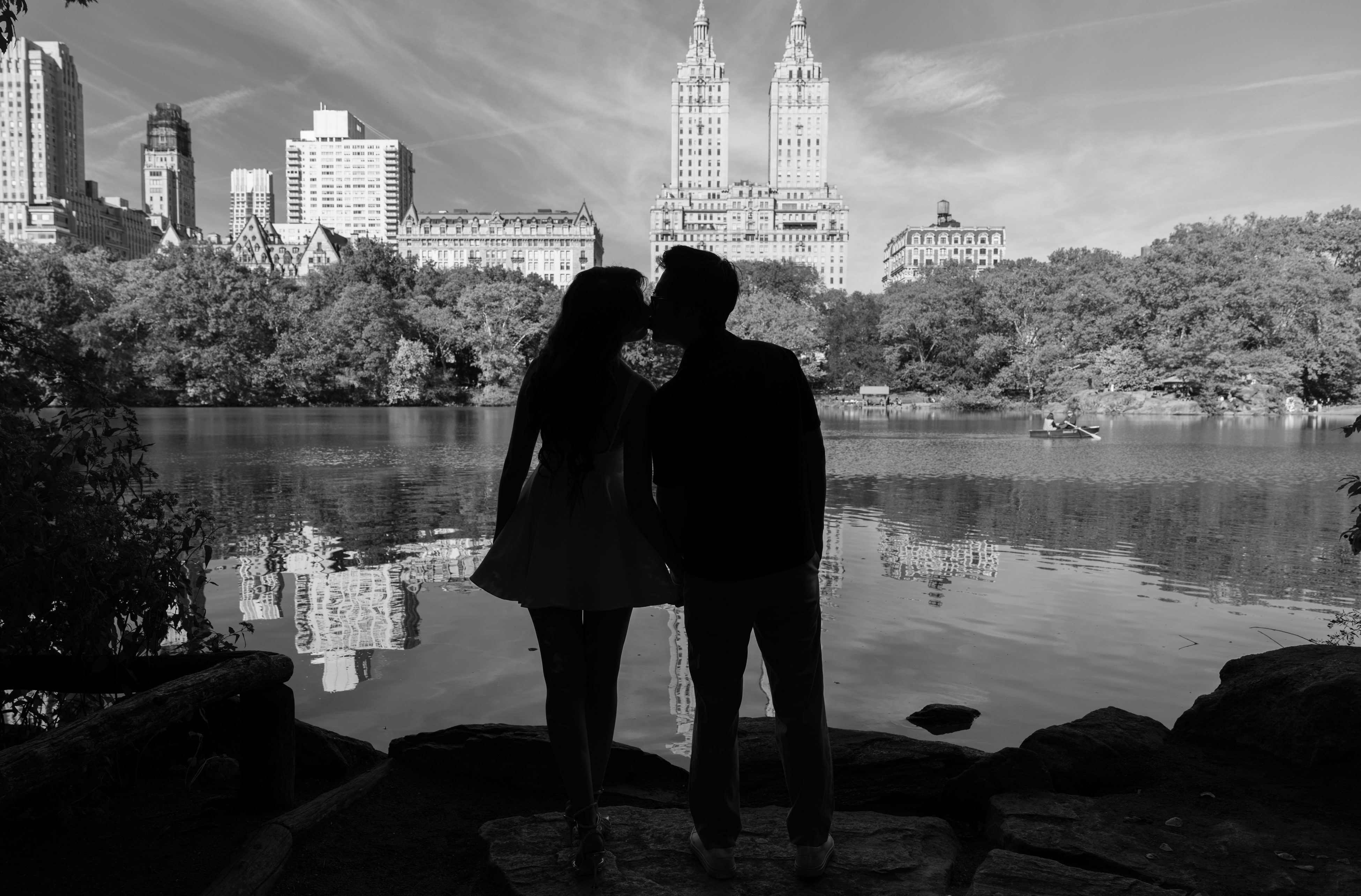 Black-and-white photograph of a couple kissing beside a lake with city buildings beyond